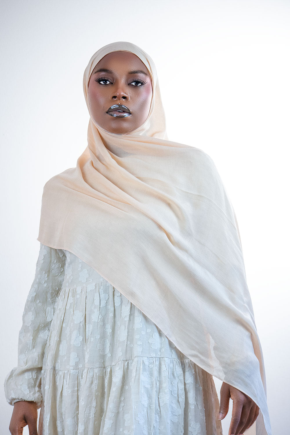 Woman wearing a beige hijab and light-colored dress against a white background